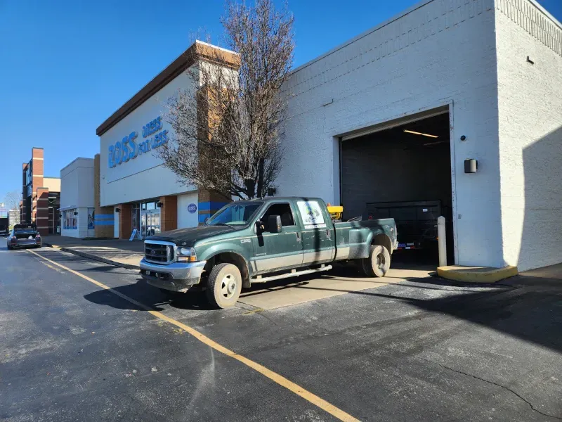 Green pickup truck parked near a white building with a garage door open. Blue sky.