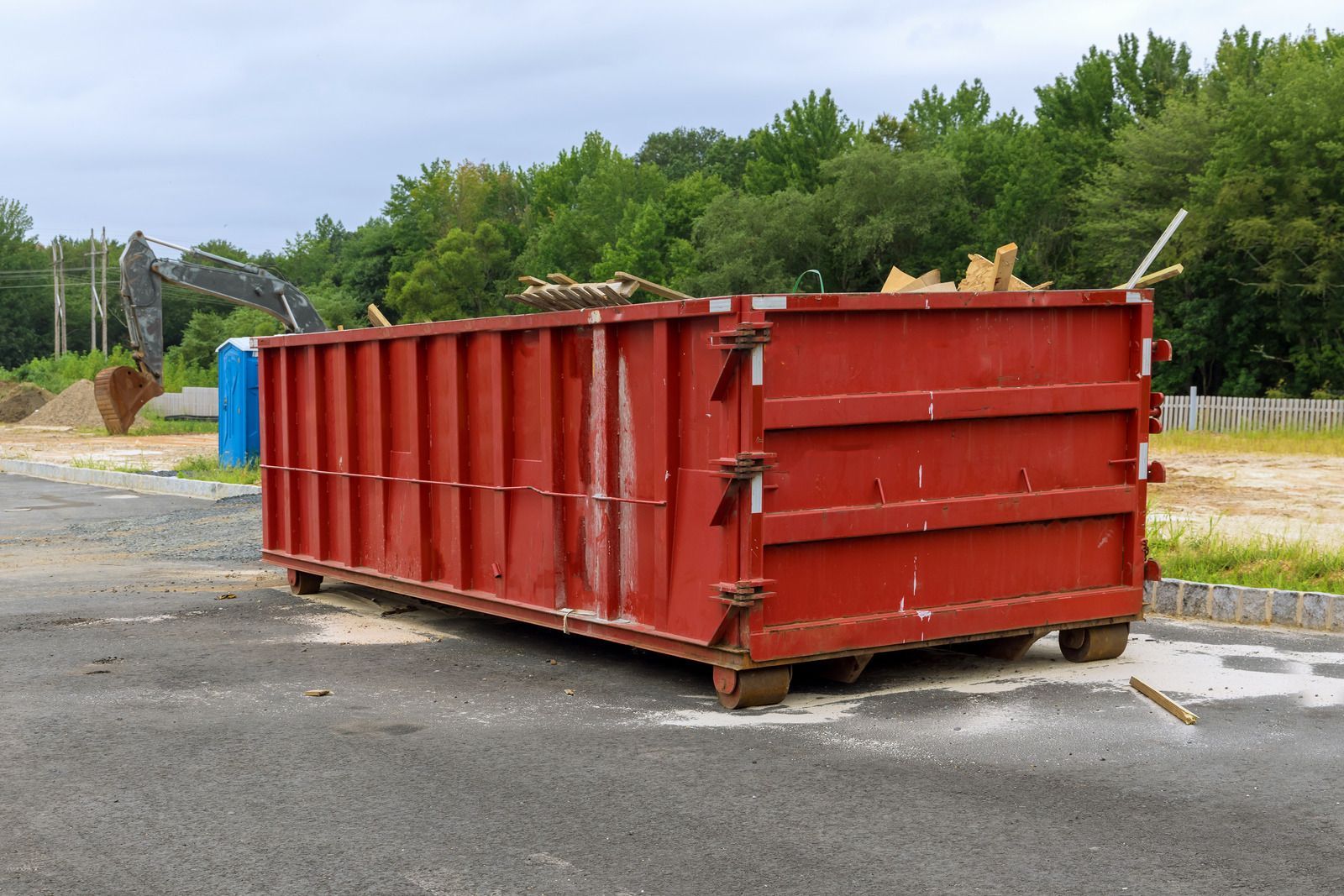 Red dumpster on wheels, partially filled with debris, sits on pavement near construction site.