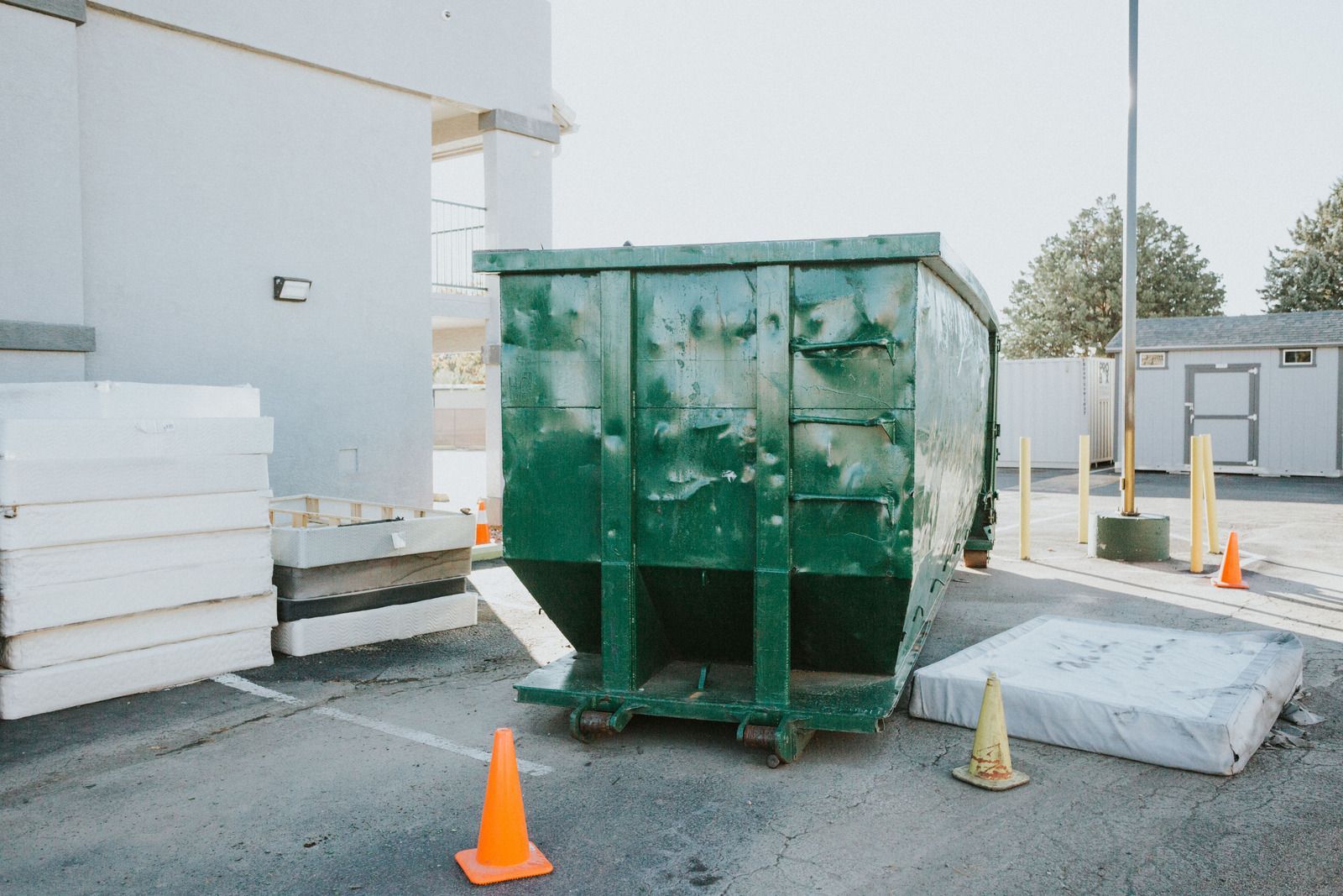 A green dumpster is sitting in a parking lot next to orange cones.