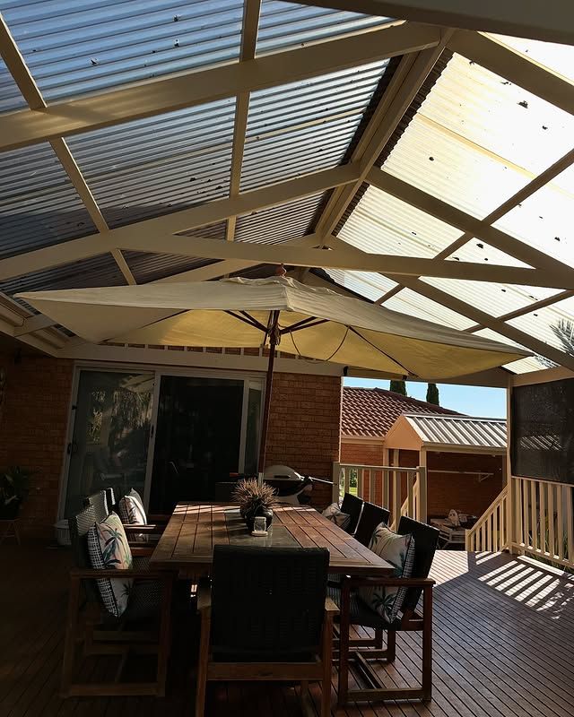 A patio with a wooden table and chairs, covered by a translucent roof. An open umbrella and barbecue are present.