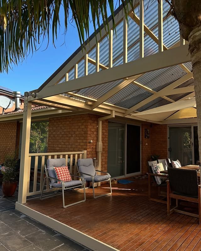 A backyard patio with a wooden deck, brick house, and a clear corrugated roof. There are chairs and a table with cushions.