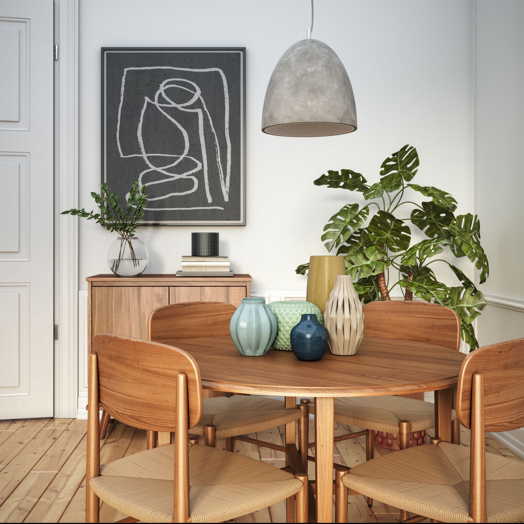 A dining room with a wooden table and chairs. There's a plant, art, and vases on the table and a pendant light overhead.