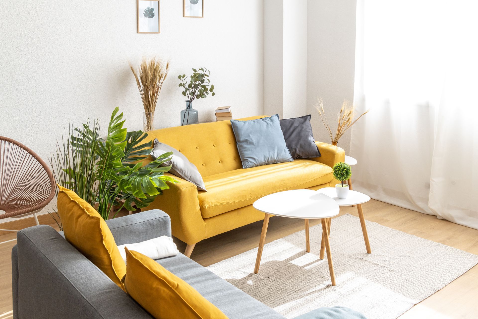 A bright living room with a yellow sofa, gray sectional, white coffee tables, and plants. Sunlight streams in from a window.