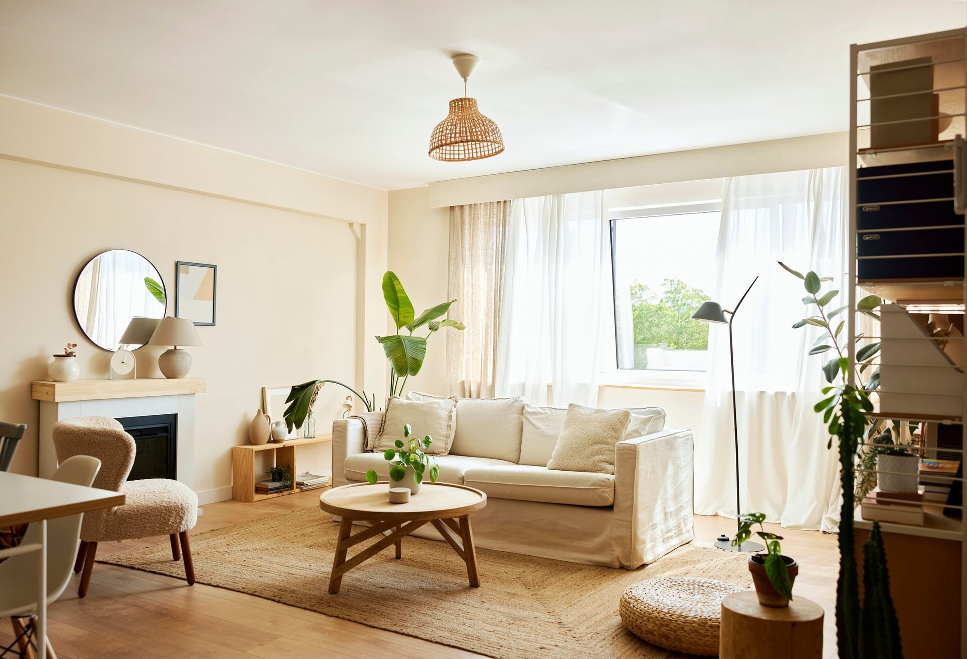 Cozy living room with a white sofa, round wooden coffee table, and large window with sheer curtains. Plants and neutral tones create a warm atmosphere.