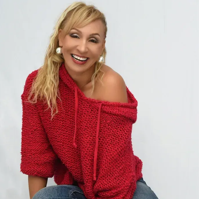 Blond woman with a big smile wearing a red off-the-shoulder sweater and jeans; seated against a white background.