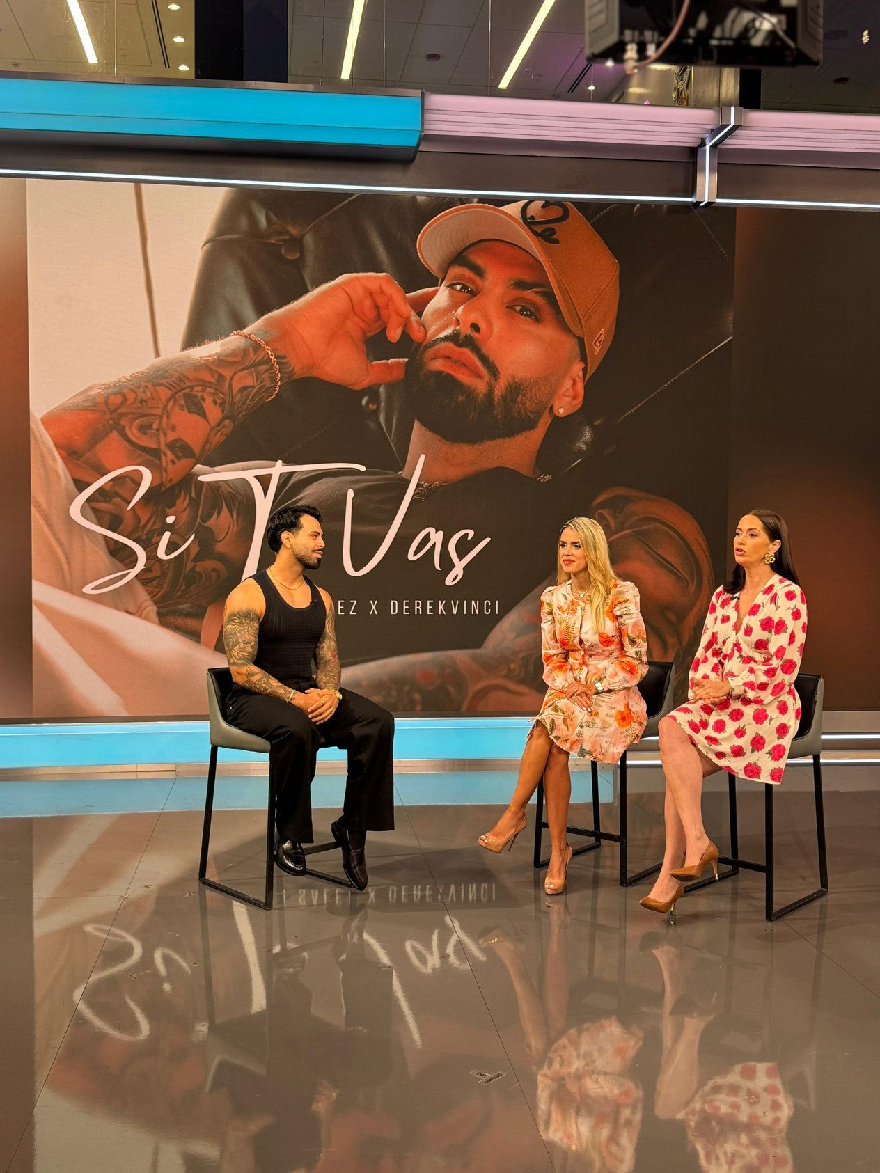 A man with tattoos sits for an interview with two women on a TV set. A backdrop shows his photo and song title 