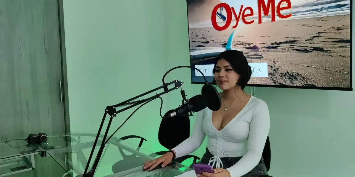 A woman with short dark hair sits in front of a microphone in a studio, holding a phone. A screen behind her reads 