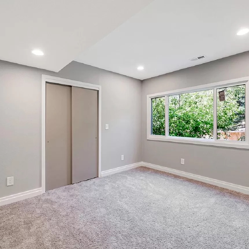 An empty bedroom with a carpeted floor , sliding doors , and two windows.