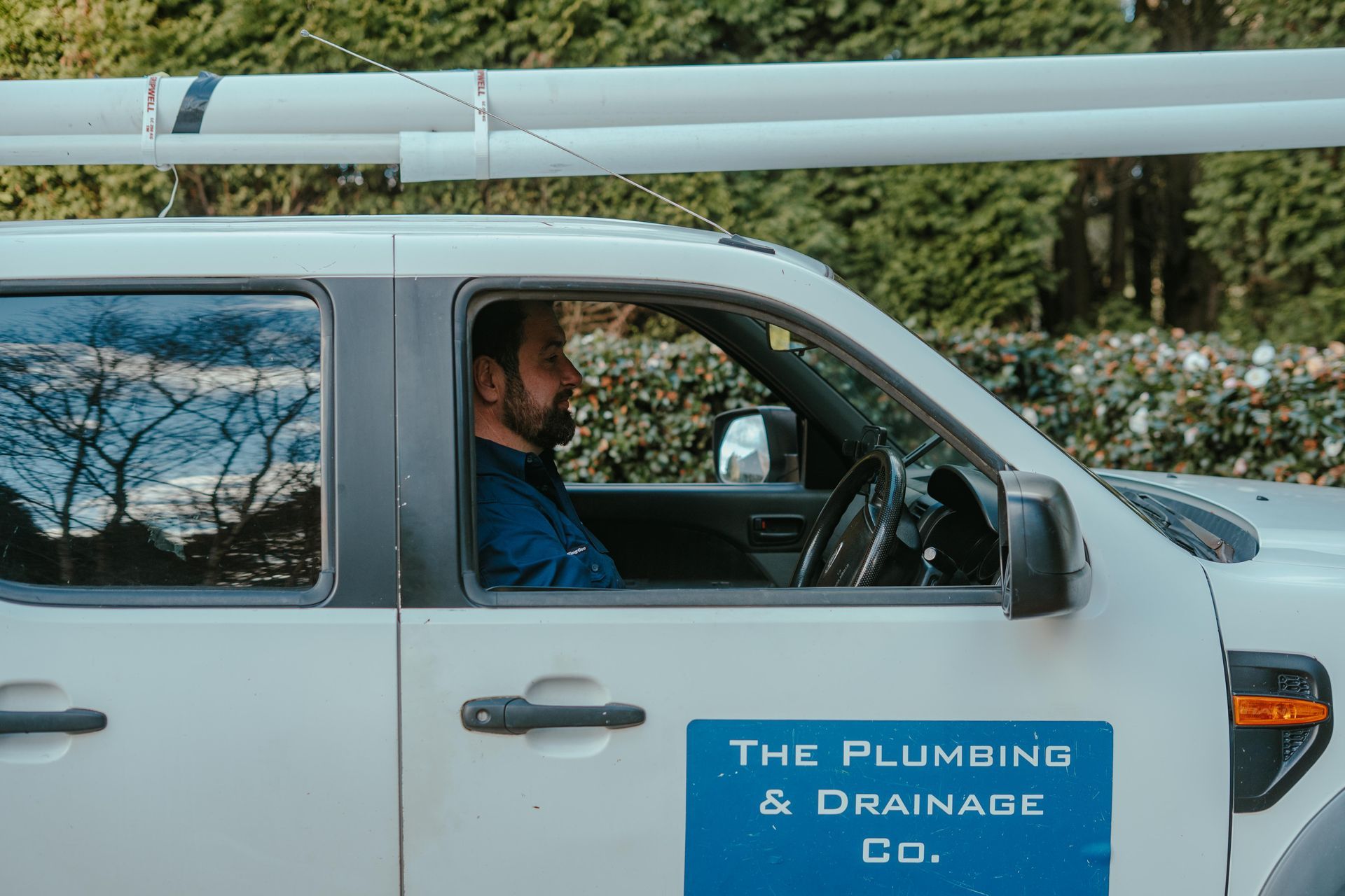 White plumbing company van with a driver, parked outdoors with pipes on the roof rack — The Plumbing & Drainage Co. in Southern Highlands, NSW