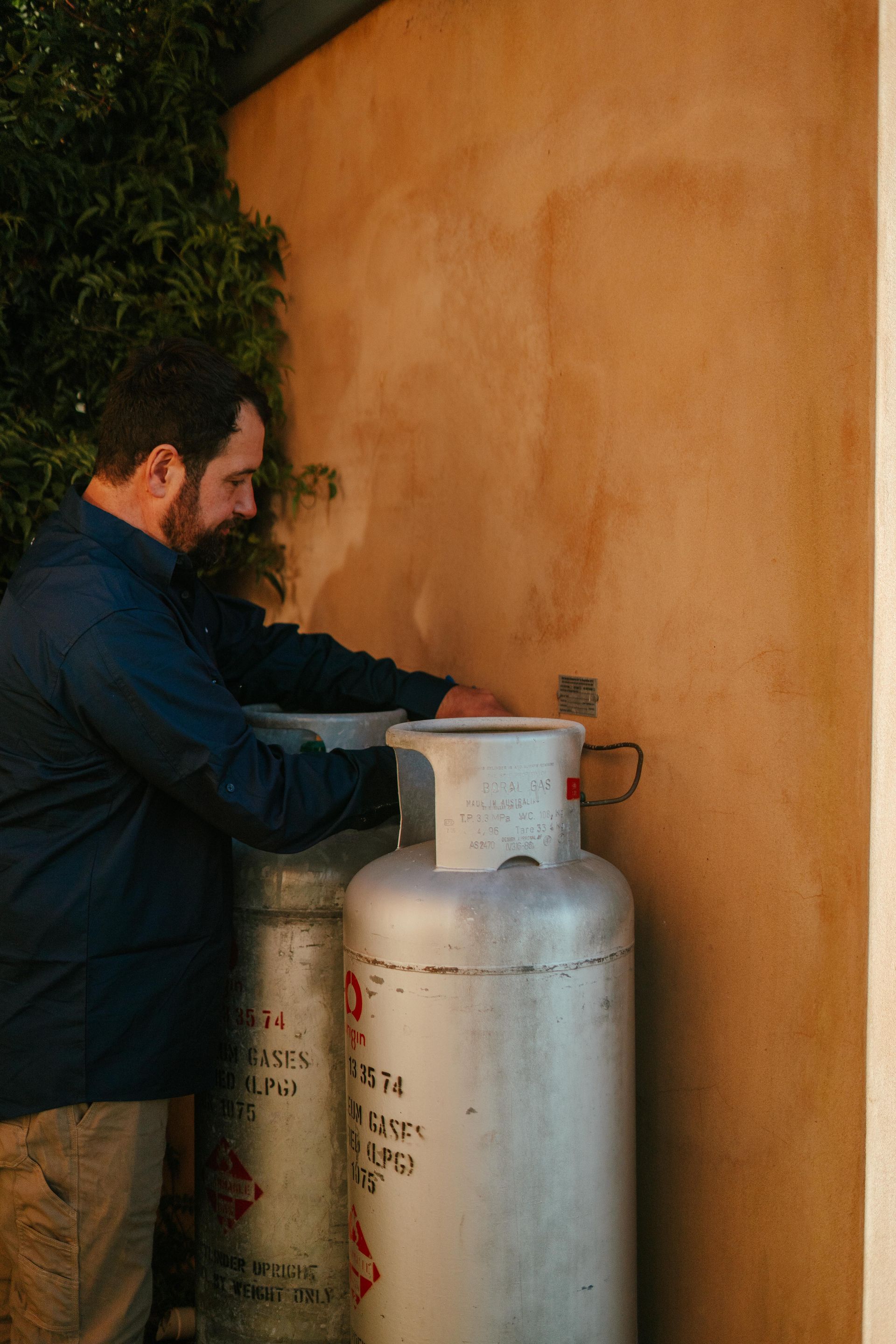 Man standing beside a silver propane tank against an orange wall outdoors — The Plumbing & Drainage Co. in Bowral, NSW