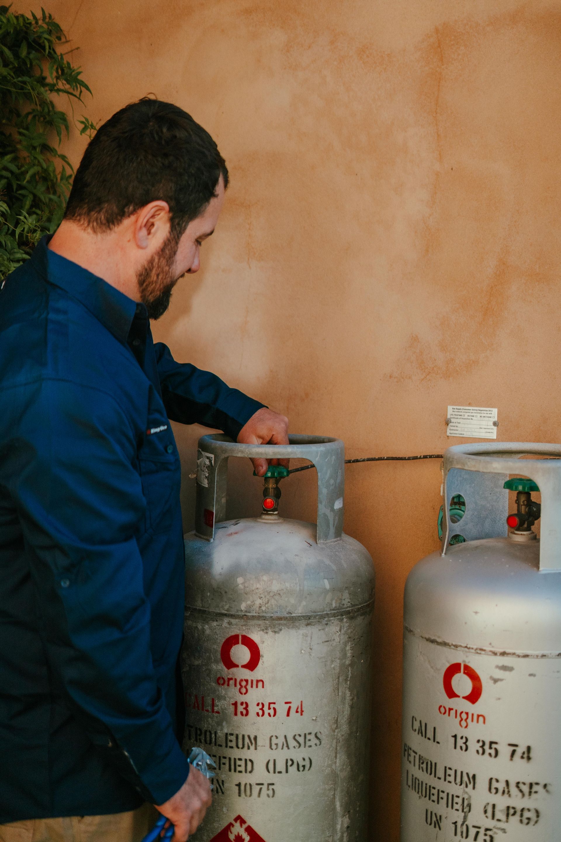 Man checking a gas cylinder valve next to another cylinder against a tan wall — The Plumbing & Drainage Co. in Campbelltown, NSW