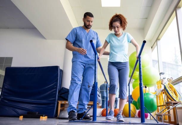 A man is helping a woman walk on parallel bars in a gym — Monroe, LA — Coleman Chiropractic Clinic