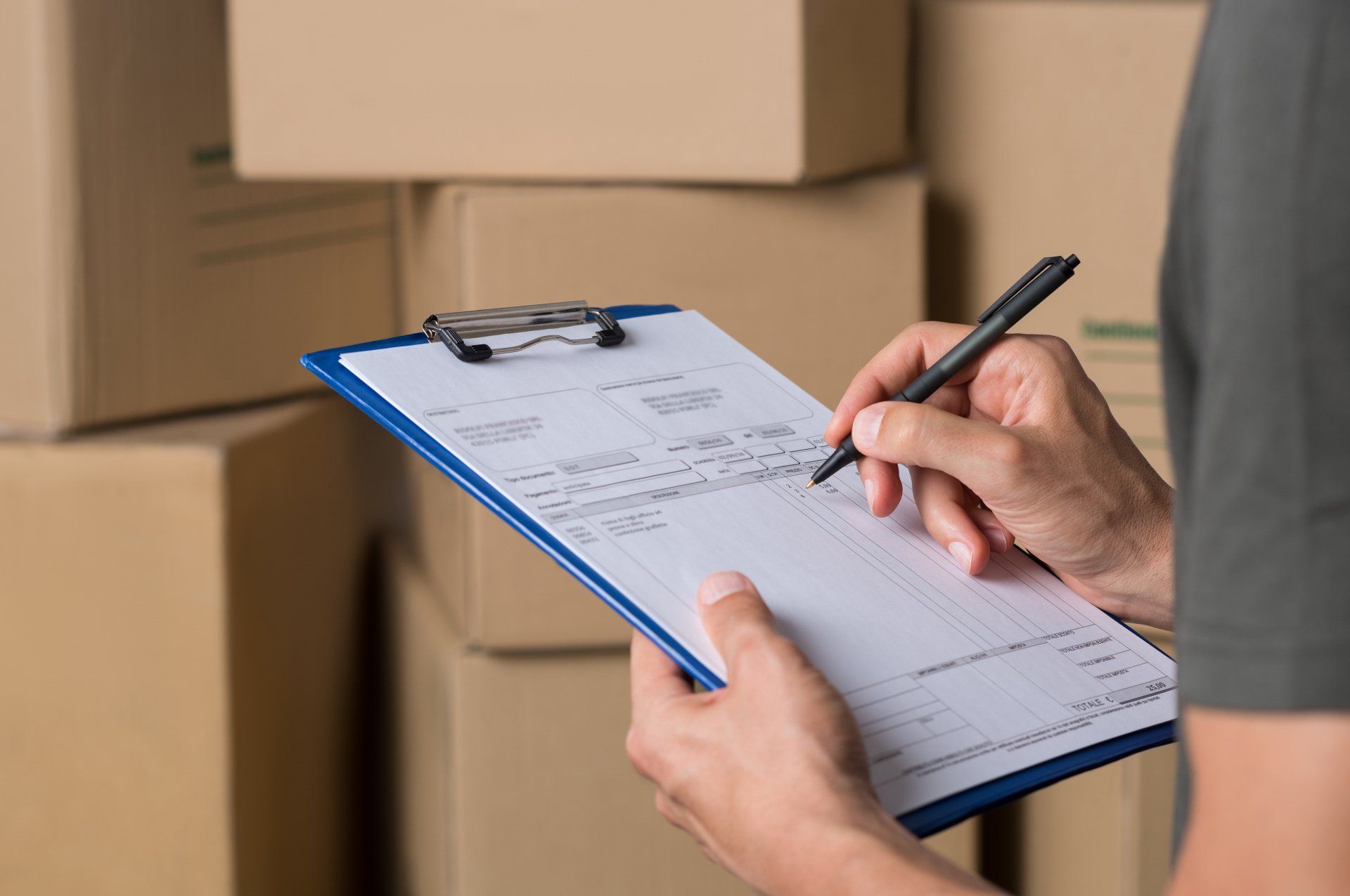 A person is writing on a clipboard in front of a pile of boxes.
