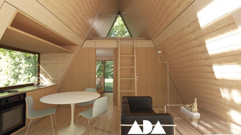 Interior of a tiny home with kitchen, desk, ladder to loft, and large windows overlooking greenery.