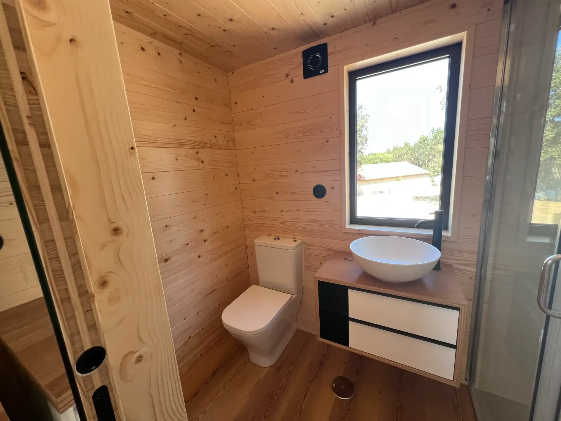 Bathroom with light wood walls, white toilet and sink, black window frame, and small cabinet.