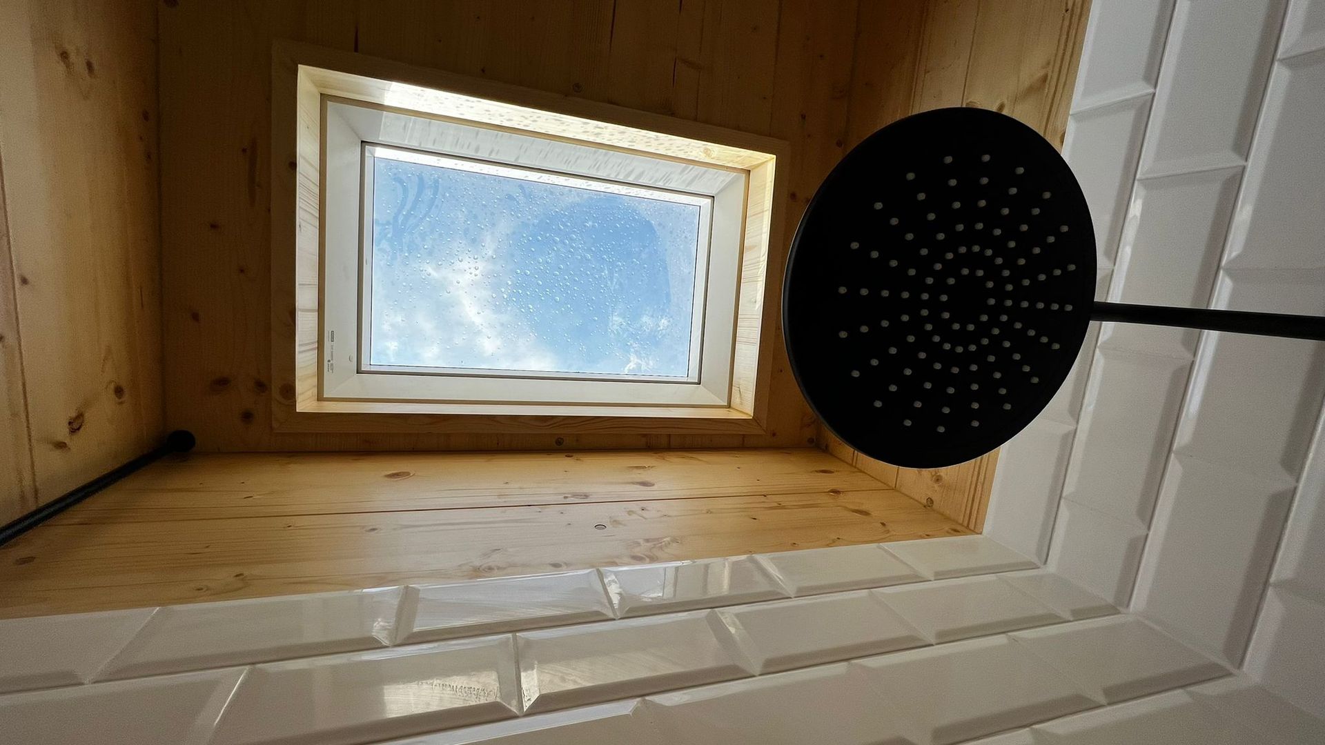 Shower with a skylight and black showerhead. White tiled wall, wood paneling, and a view of the blue sky.