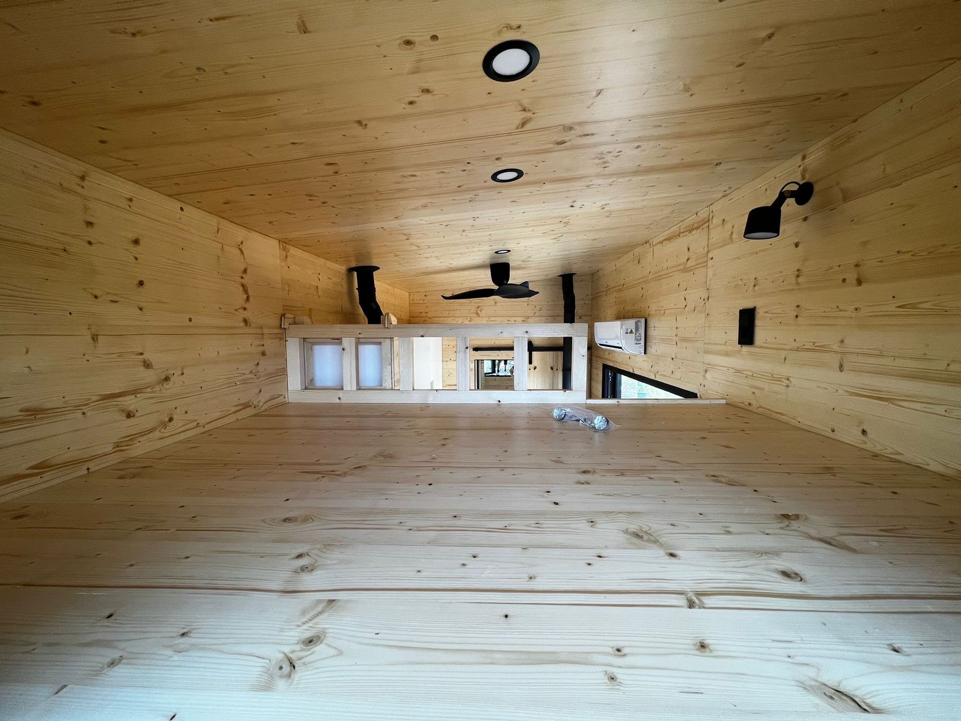 Interior view of a wooden tiny home with a loft, showing the floor, walls, and ceiling with recessed lighting.