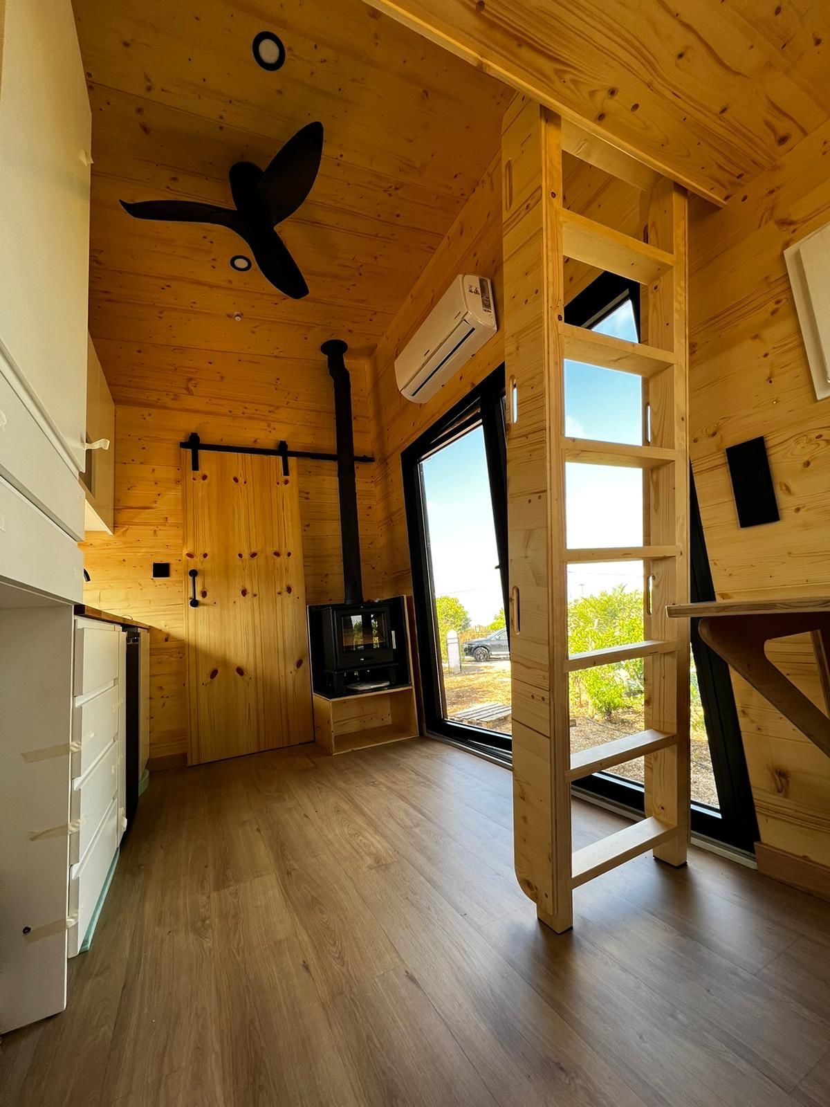 Interior of a tiny home with wood paneling, a ladder to a loft, fireplace, and large window.