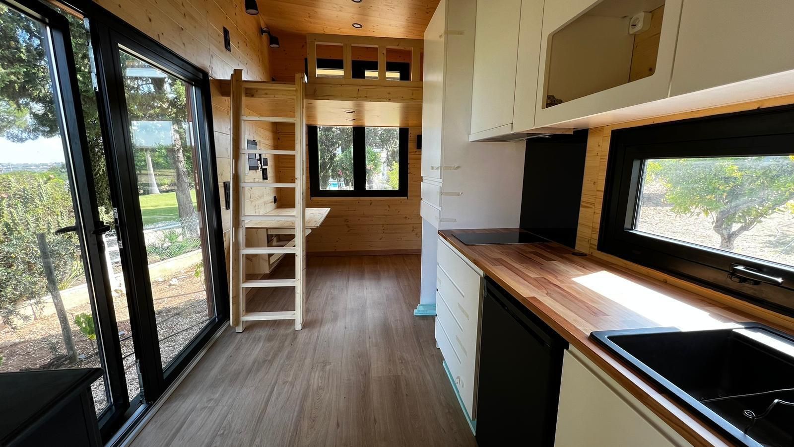 Interior of a tiny house with kitchen, loft, desk, and sliding glass door to outside view.