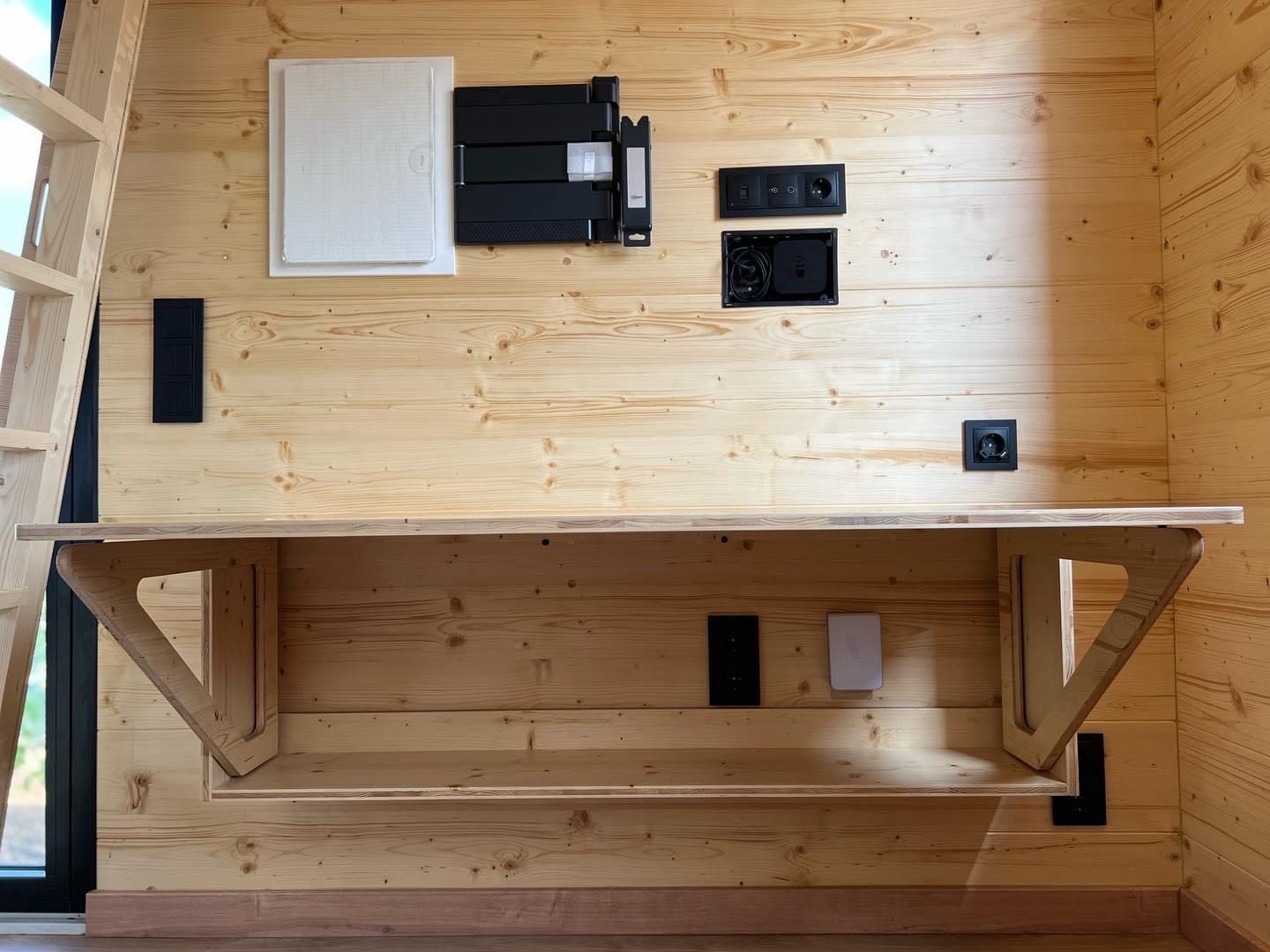 Wooden desk mounted on a wooden wall, with outlets, a ladder, and a window visible.