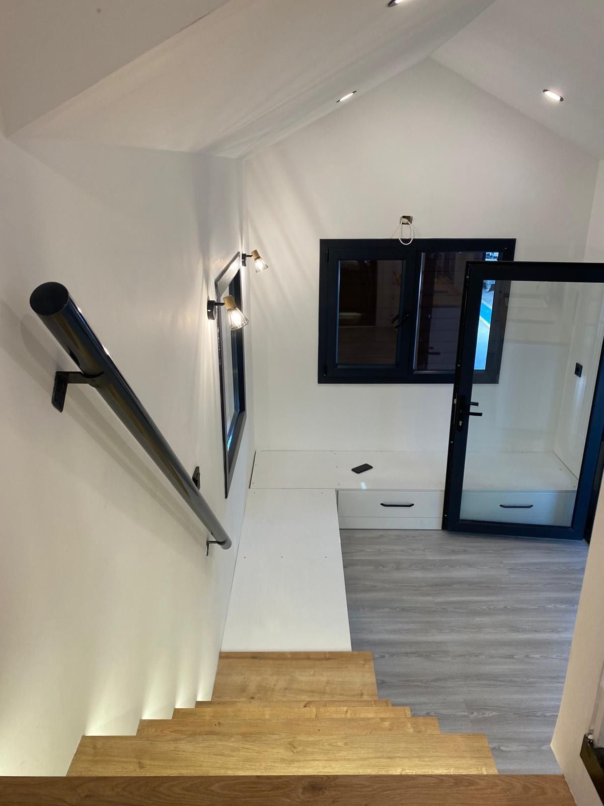 Staircase with wooden steps, white walls, black railing, window, and door.