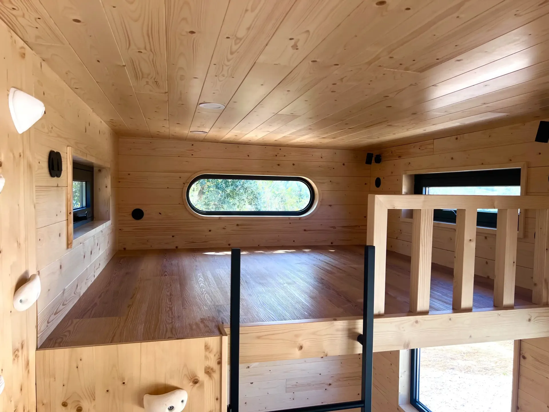 Interior of a small wooden treehouse with a loft, porthole window, and climbing holds on the wall.