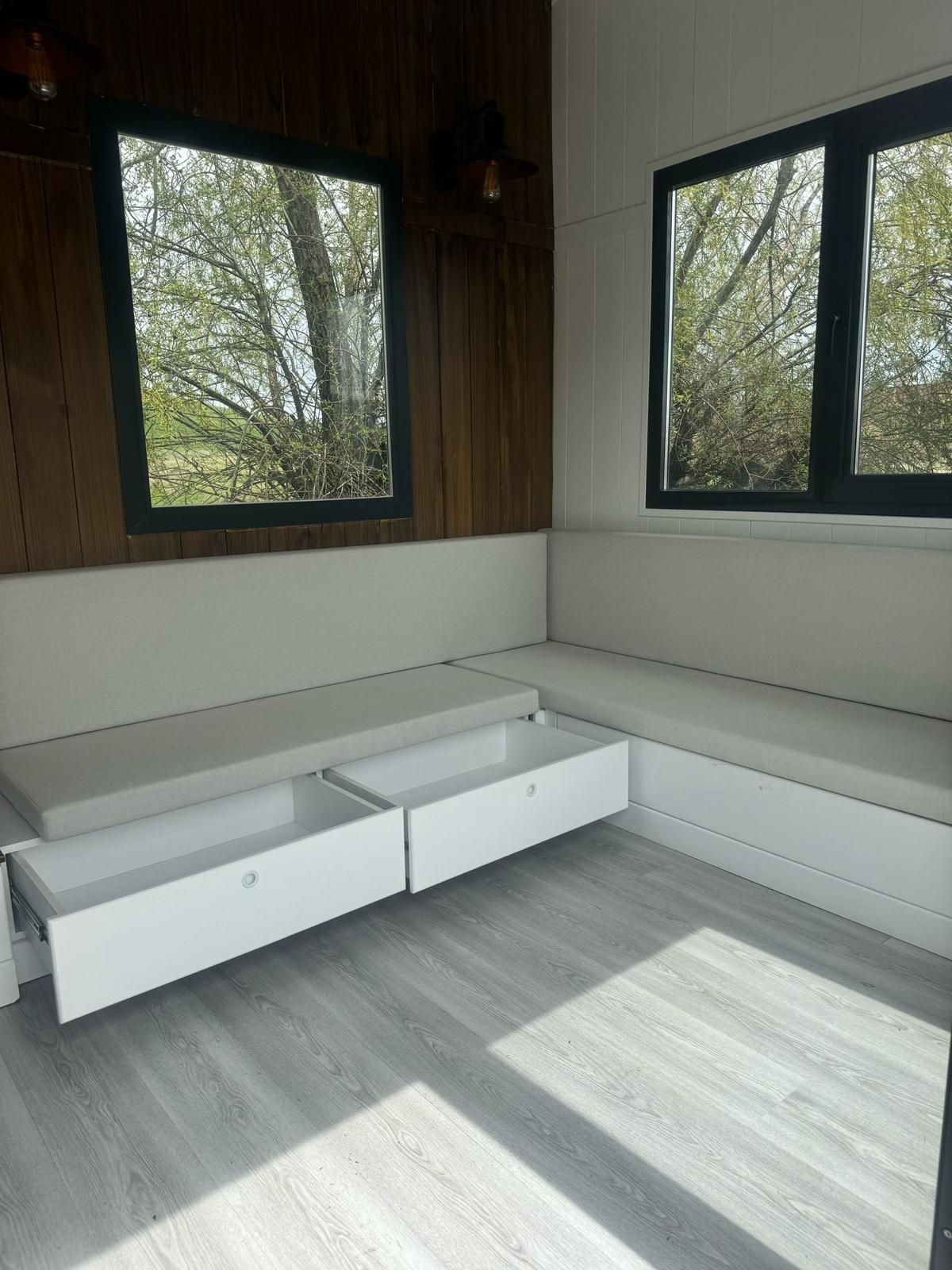 Corner seating with drawers, white cushions, two windows, wood paneling, and light gray floor.