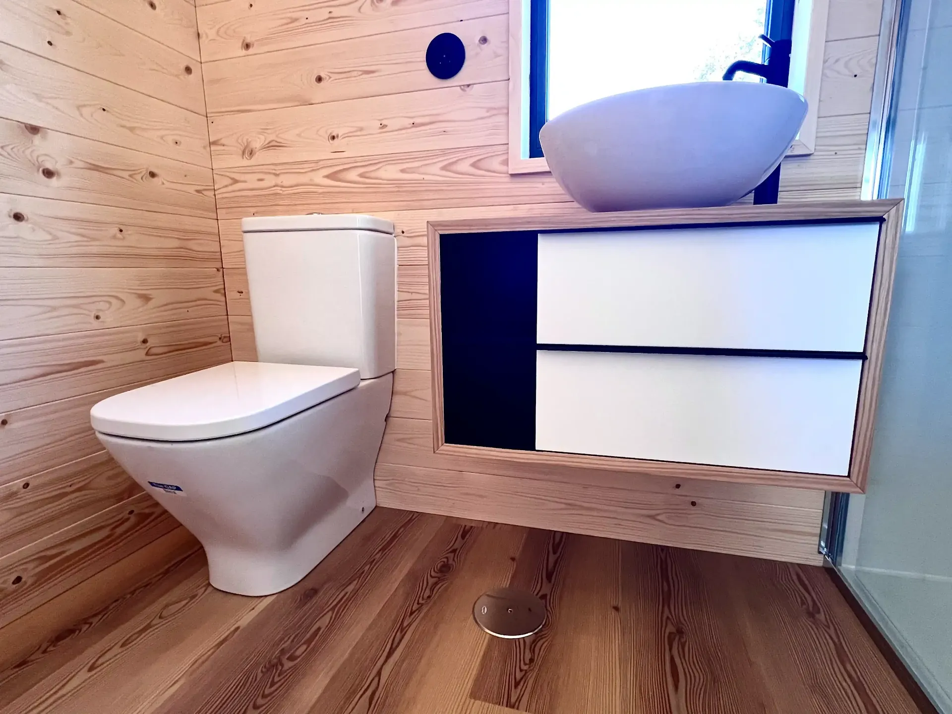 Bathroom with white toilet, vanity, and sink; wood paneling, small window.
