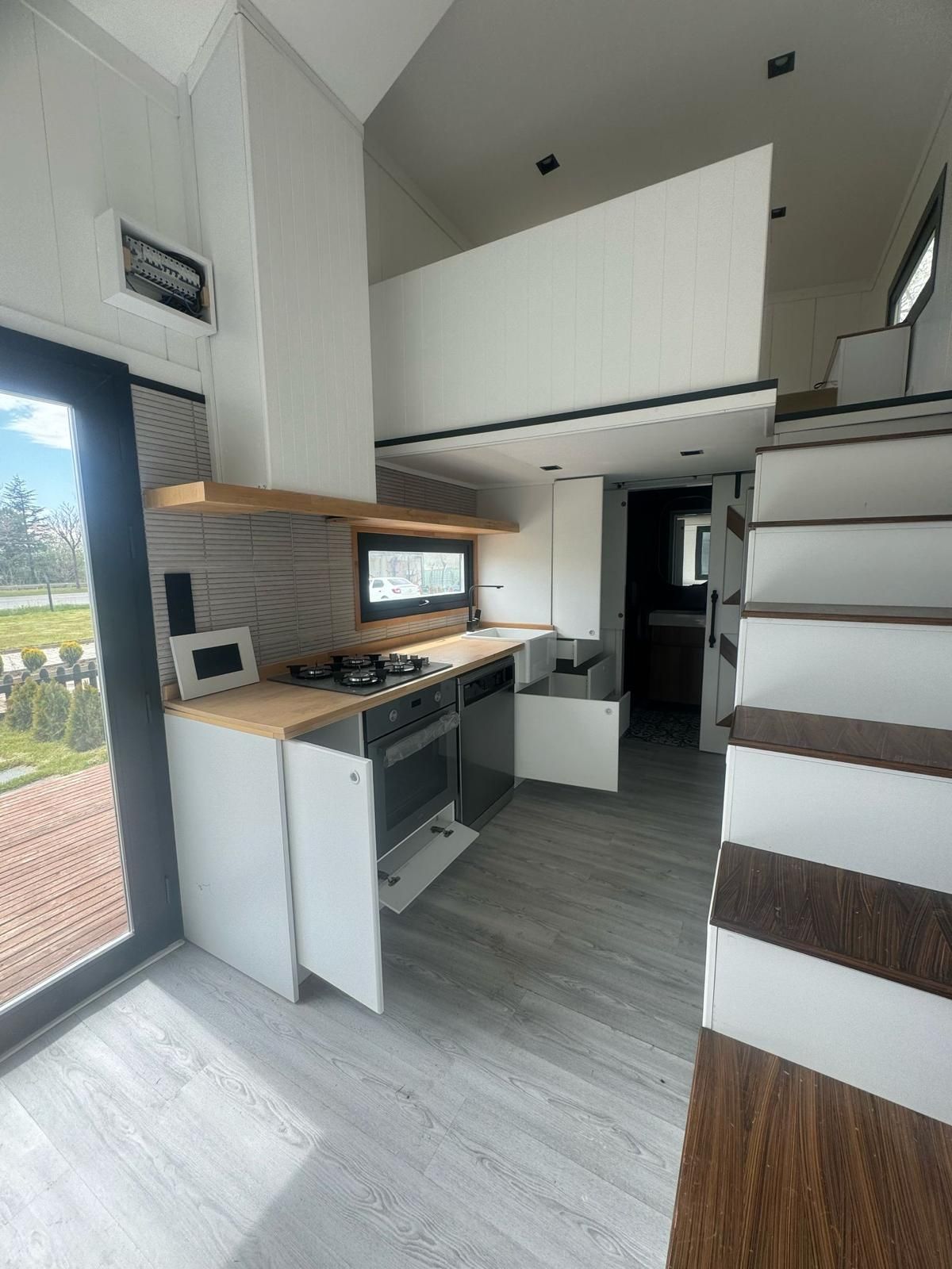 Tiny home interior with kitchen and loft, natural light, white cabinets, and wood accents.