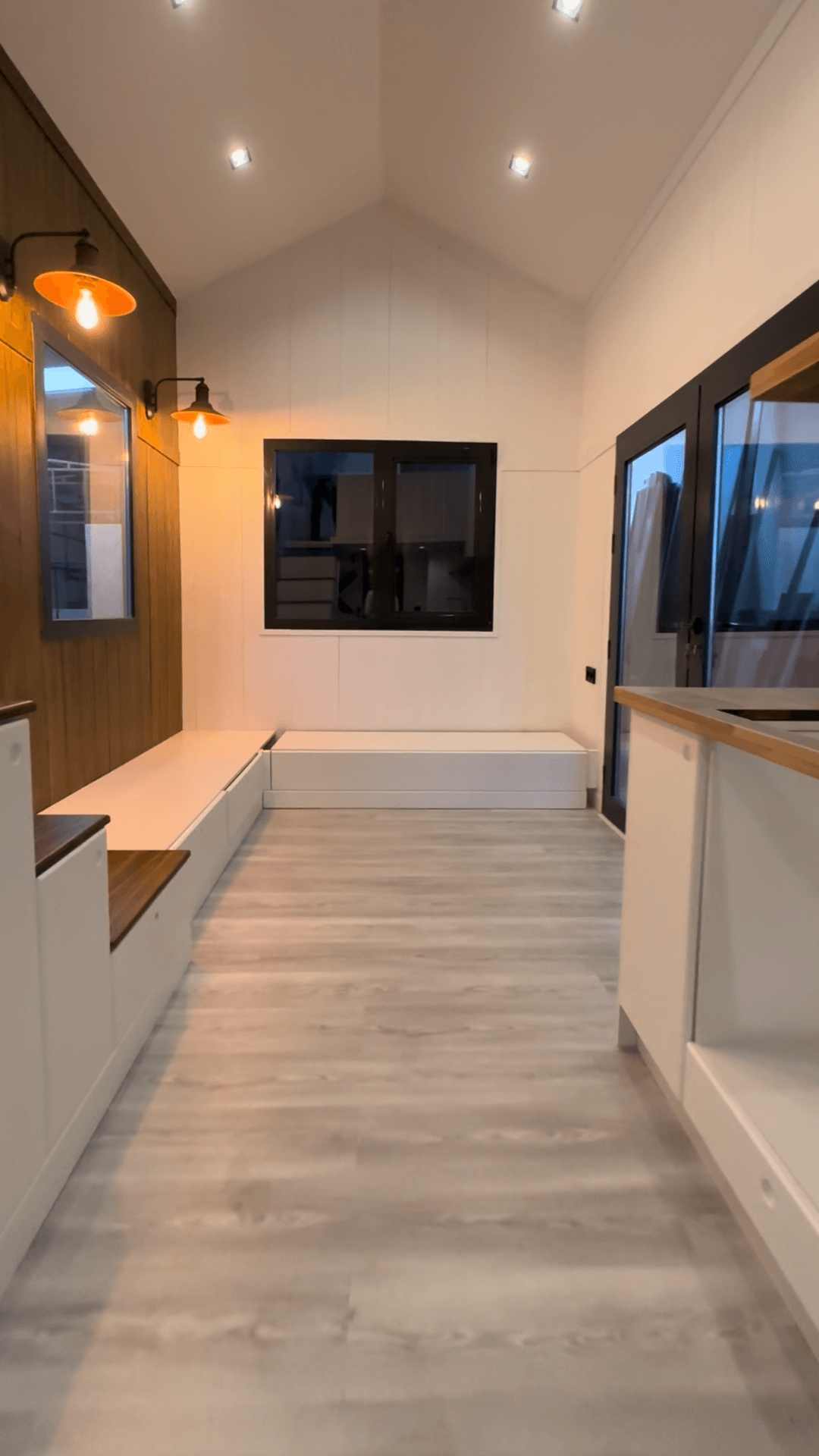Interior of a tiny home with light wood floors, white walls, built-in bench seating, and a mounted TV.
