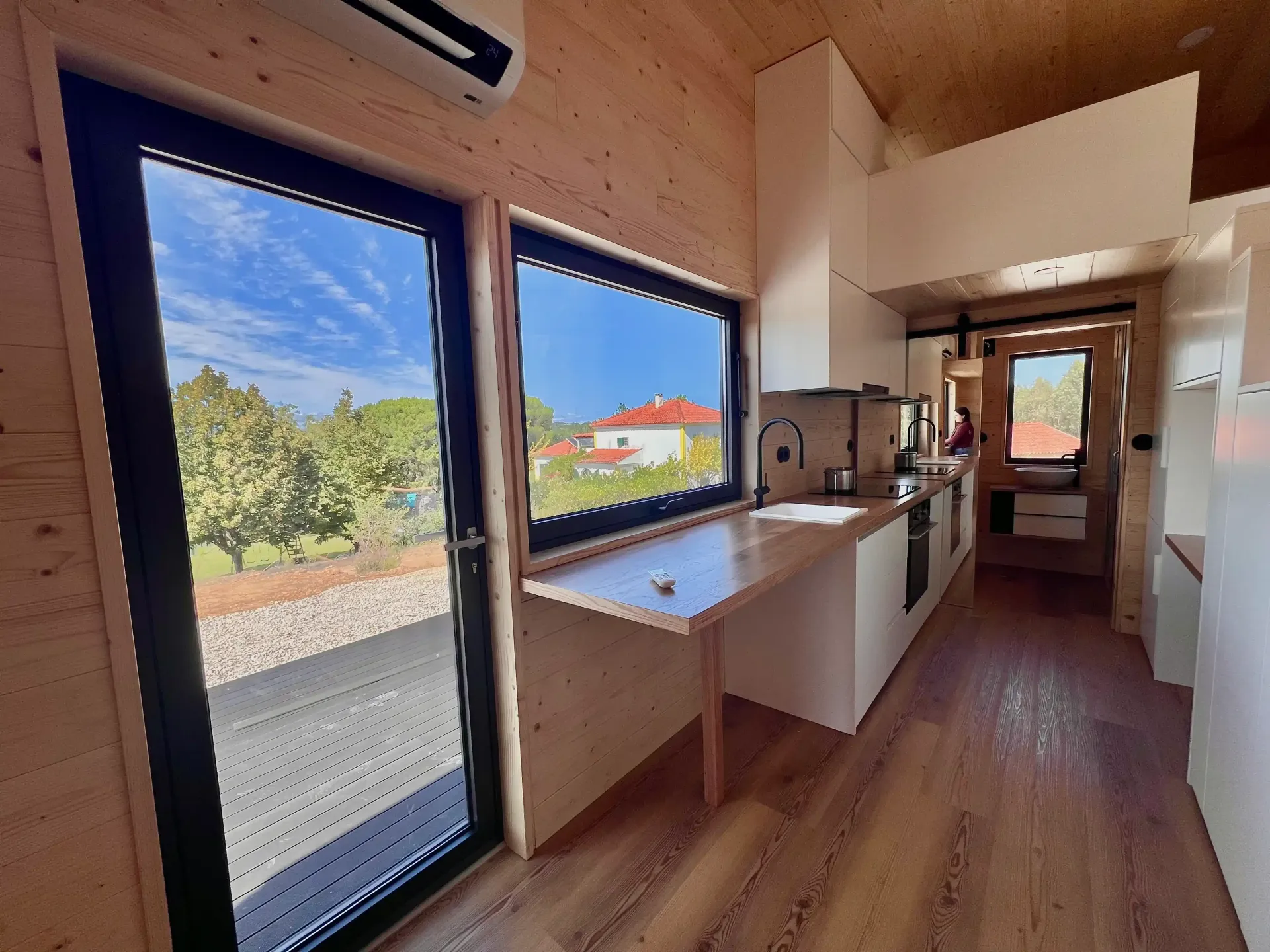 Interior of a tiny house: wooden walls, large windows with blue sky view, kitchen, and fold-down table.
