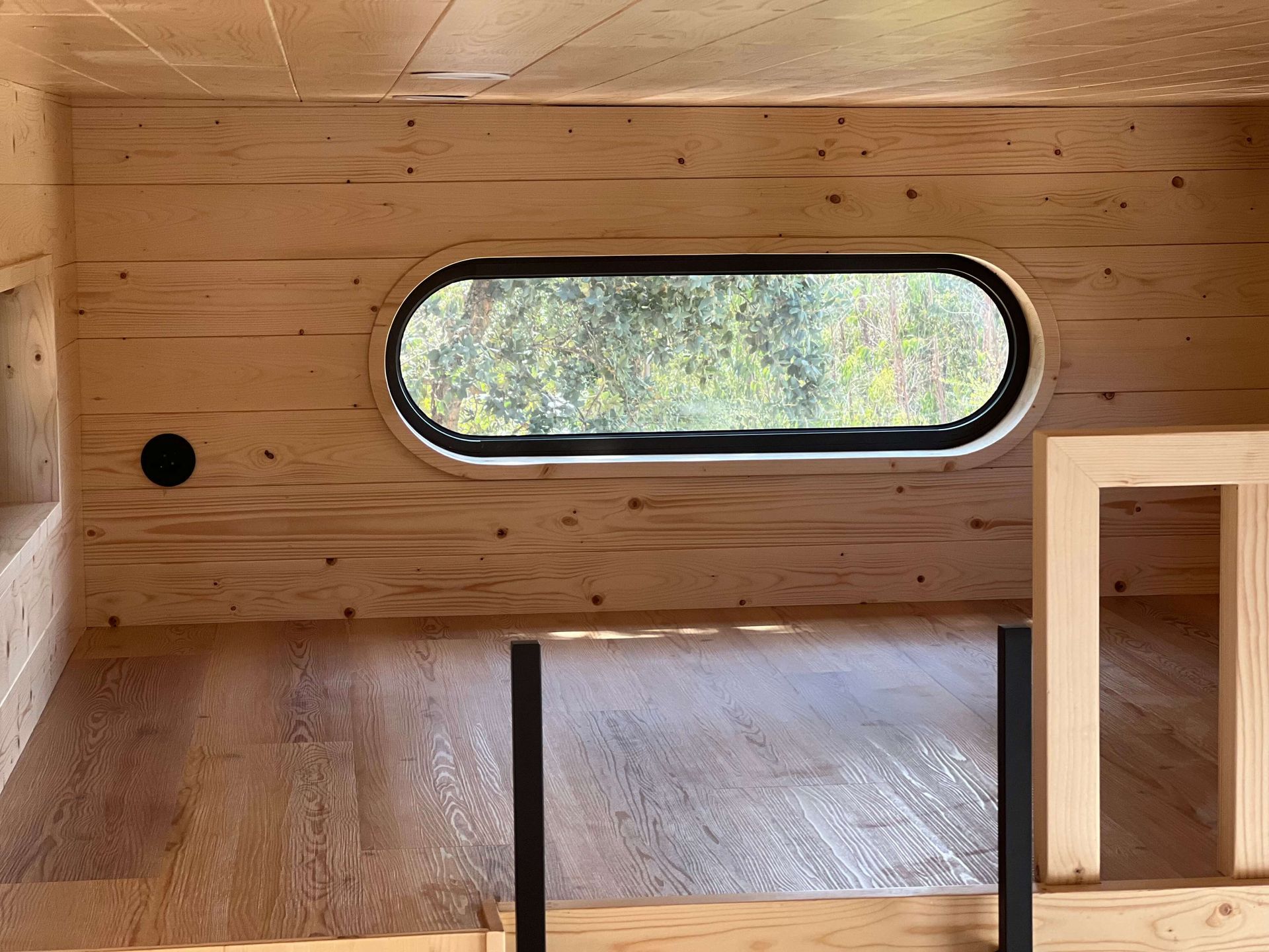 Interior view of a small wooden structure with an oval window looking out to greenery.