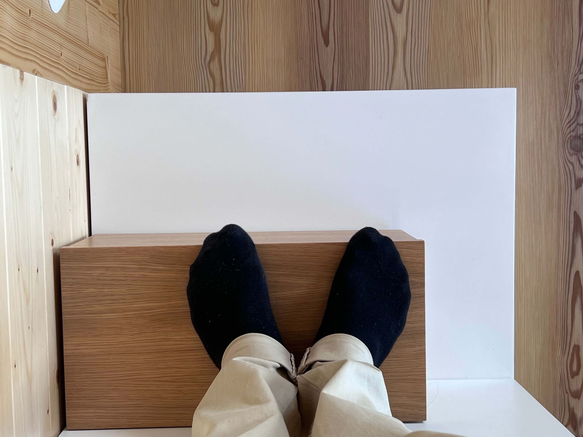 Feet in black socks, khaki pants, on a wooden box against a white backdrop in a wooden-paneled room.