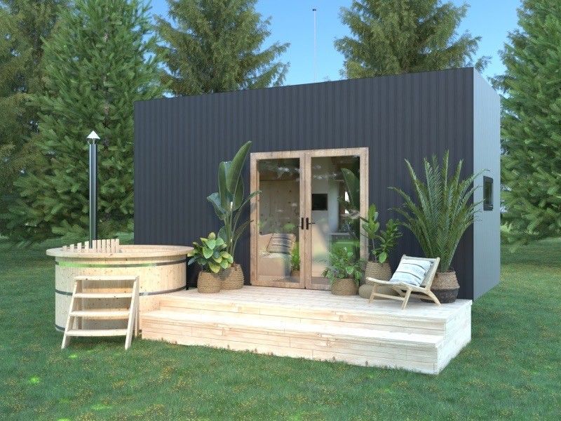 Black cabin with a wooden hot tub, surrounded by plants, on a wooden deck in a grassy area.