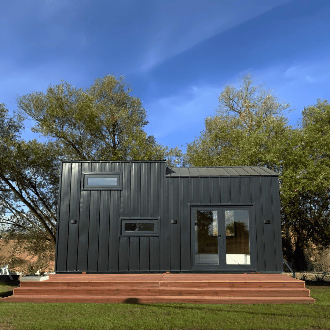 Tiny dark modern house with two windows and a glass door on a wooden deck under a blue sky.