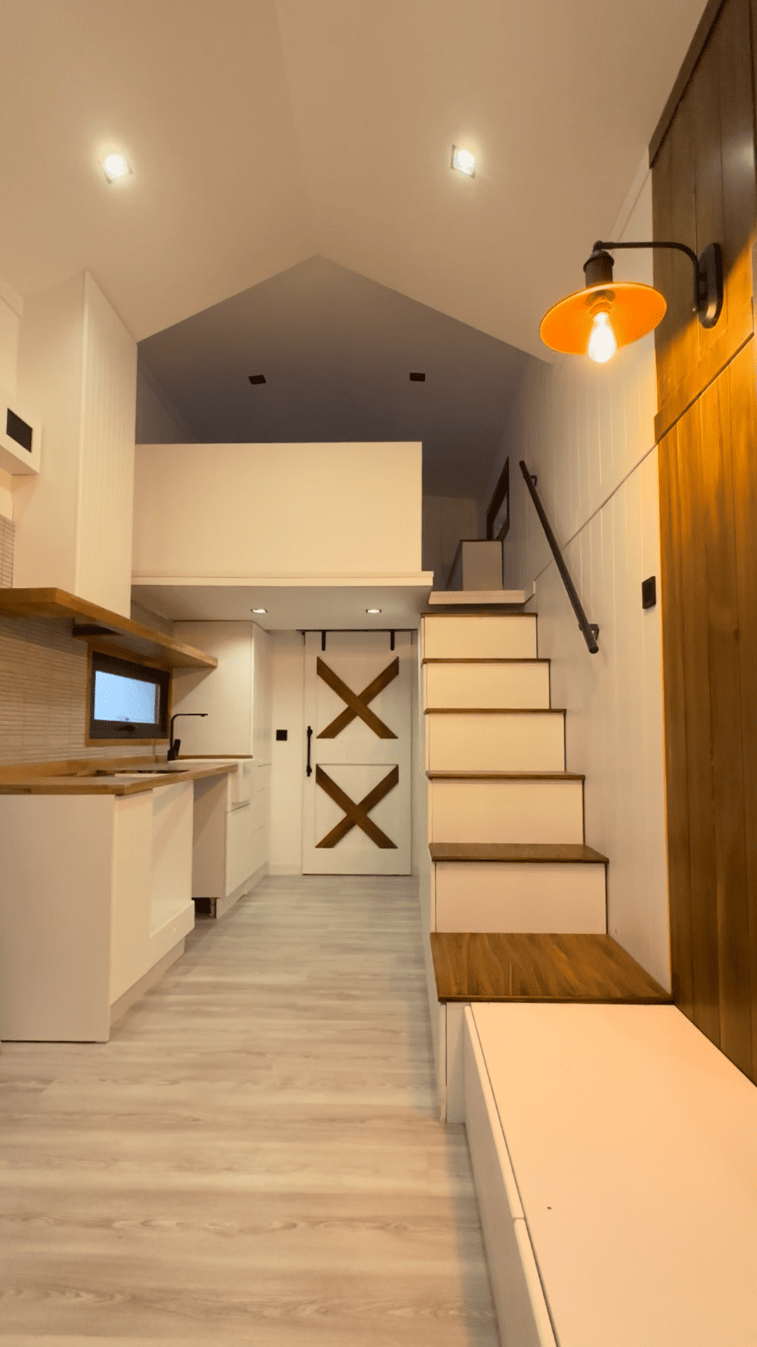 Tiny home interior: white kitchen, loft, wooden stairs, and a built-in bench.