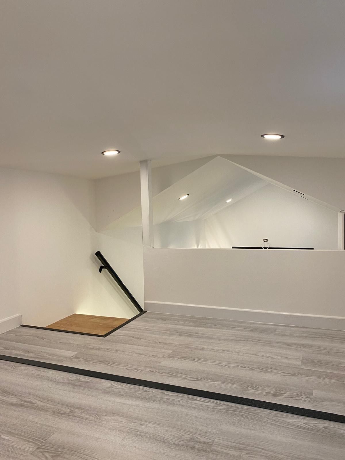 Empty, modern room with white walls, recessed lighting, and a partial second level with a glass barrier.
