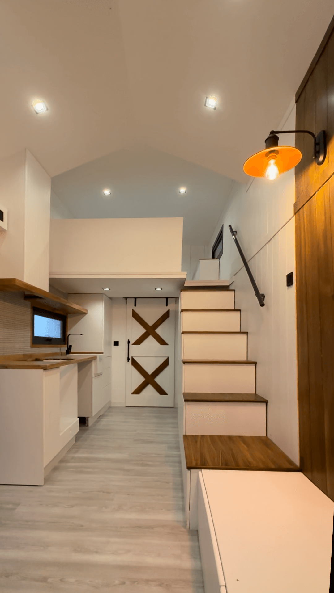 Interior of a tiny house with a kitchen, staircase, loft, and barn door. Bright white and wood finishes.
