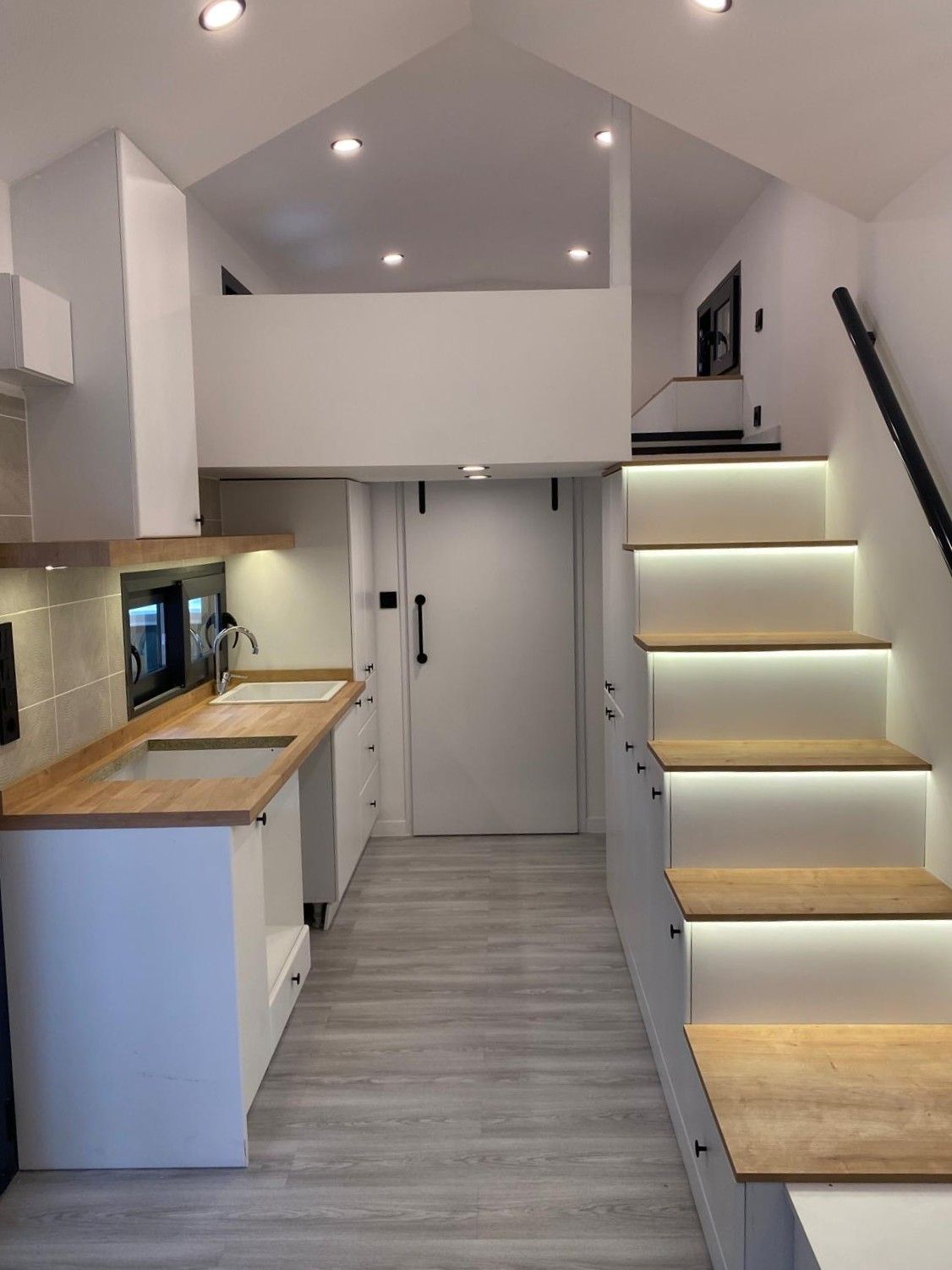 Interior of a tiny home with a kitchen and staircase leading to a loft area; white, wood tones, and LED lighting.
