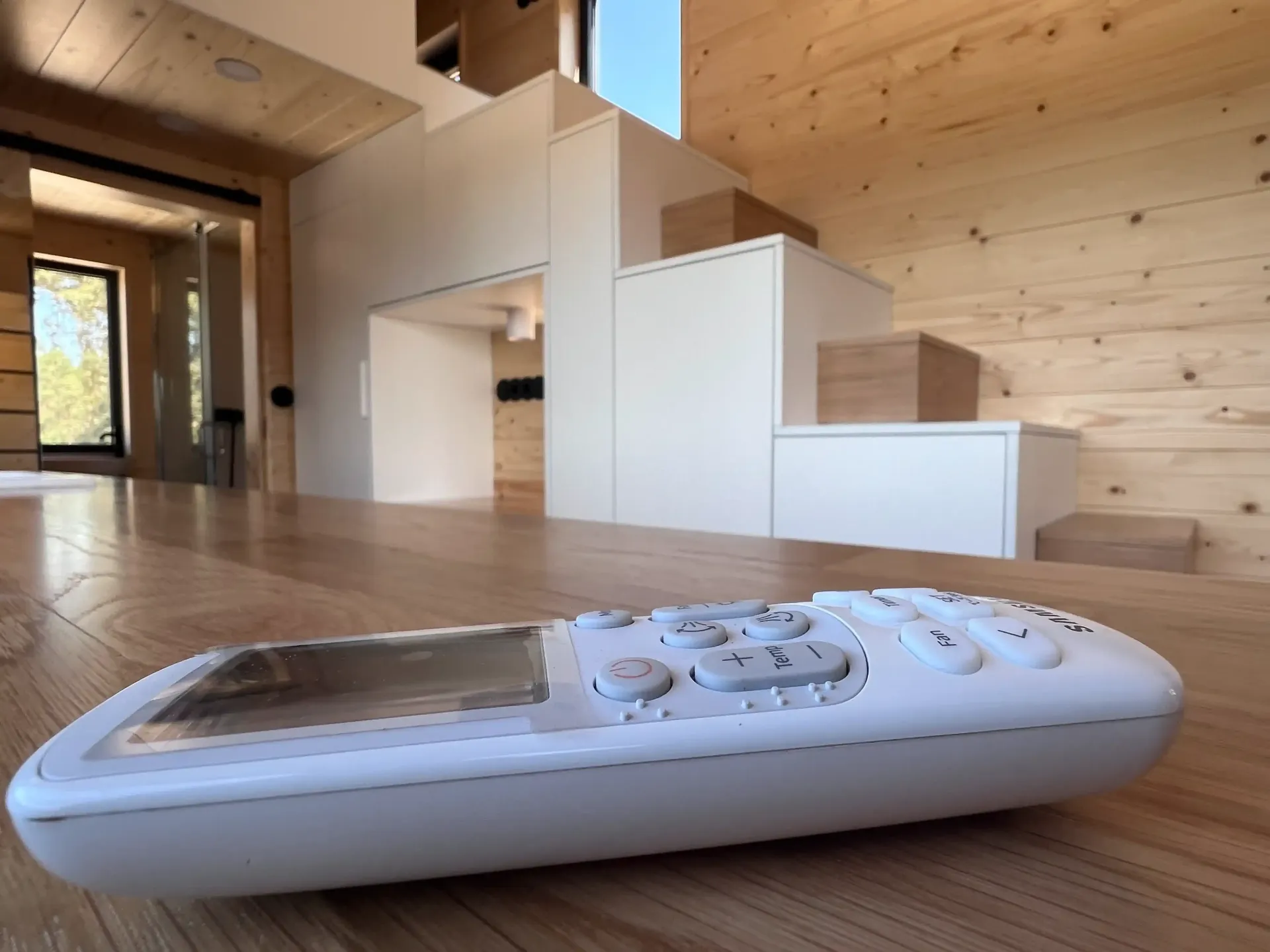 White remote control on a wooden floor, with a modern interior featuring stairs in the background.