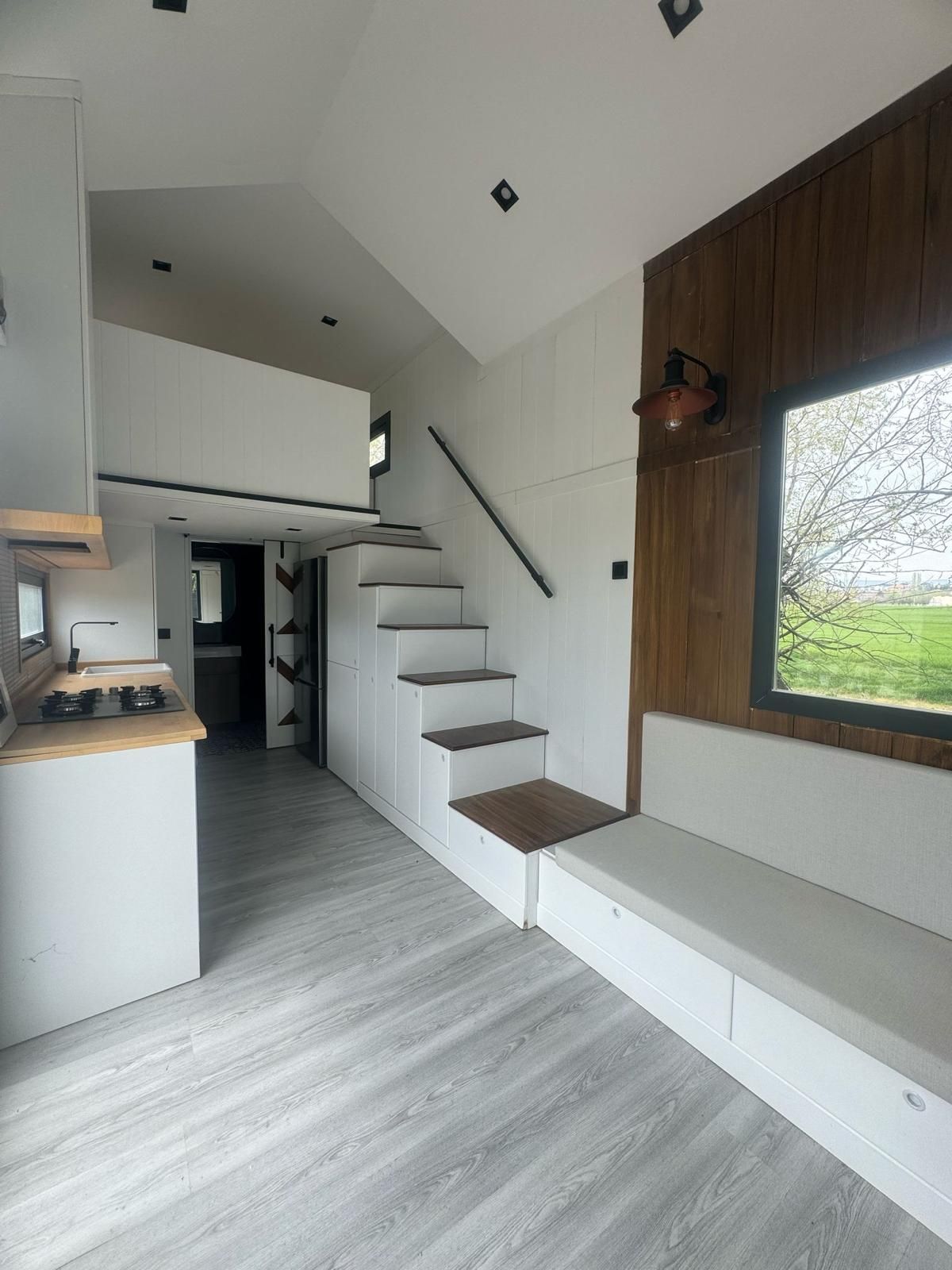 Interior view of a modern tiny house. White stairs lead to a loft. Light gray flooring, white walls, and a wooden accent wall.