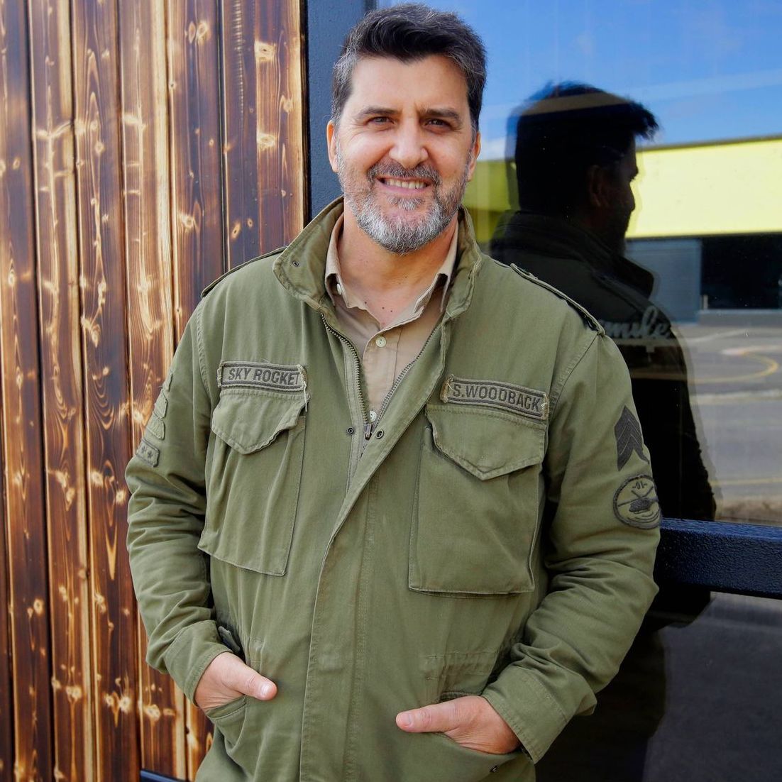 Man with graying beard in green jacket, smiling, hands in pockets, near wooden building.
