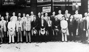 Group of men outside a building, many holding hats. Building has a sign for 