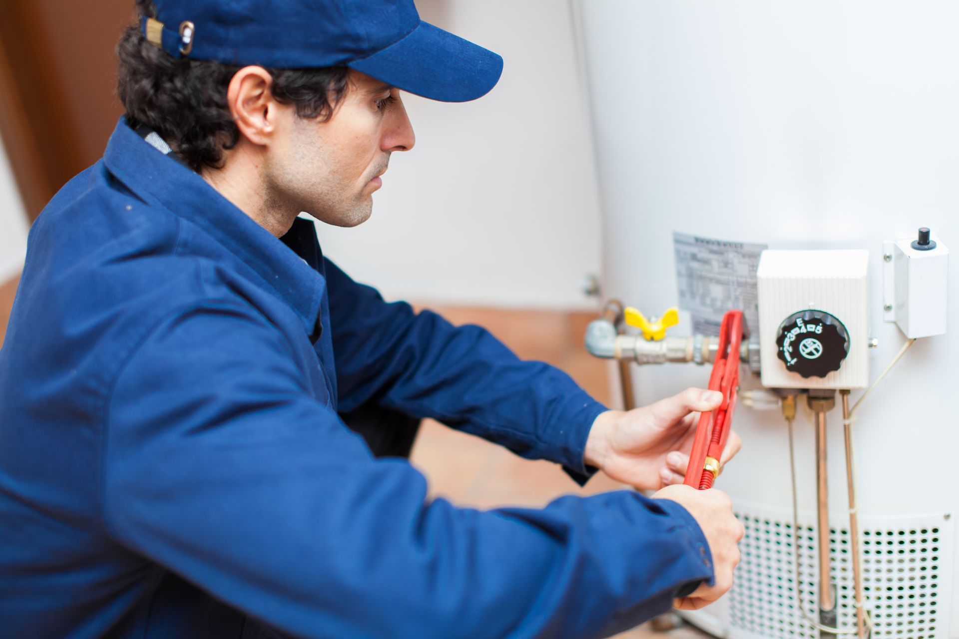 Plumber in blue uniform uses a wrench to work on a white water heater in a room.