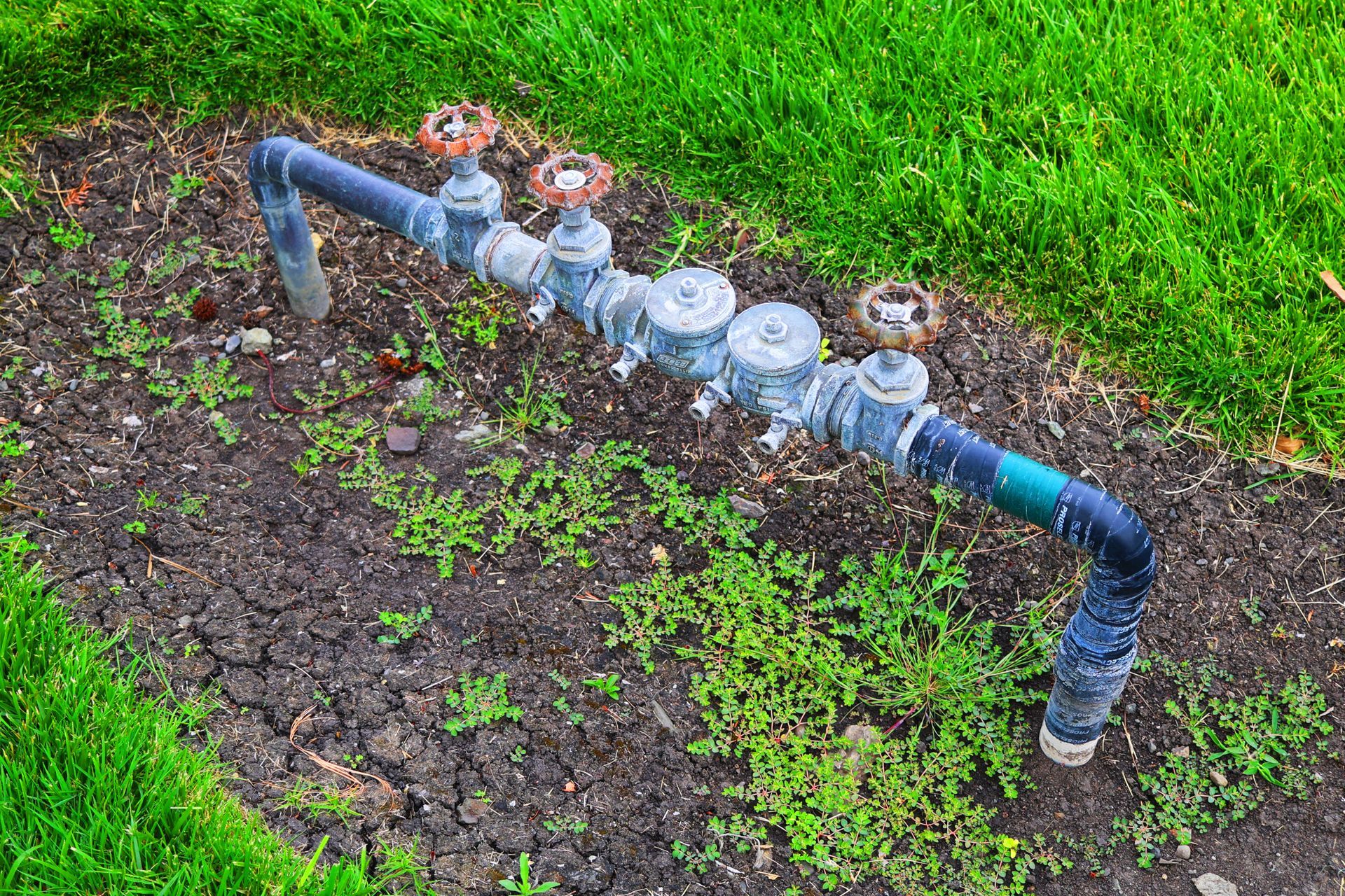 Pipes with valves for irrigation system in a garden bed with grass.