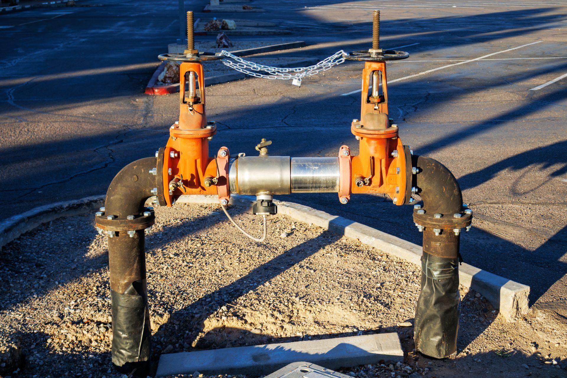 Orange valves and pipes in gravel, asphalt background, chained handles.