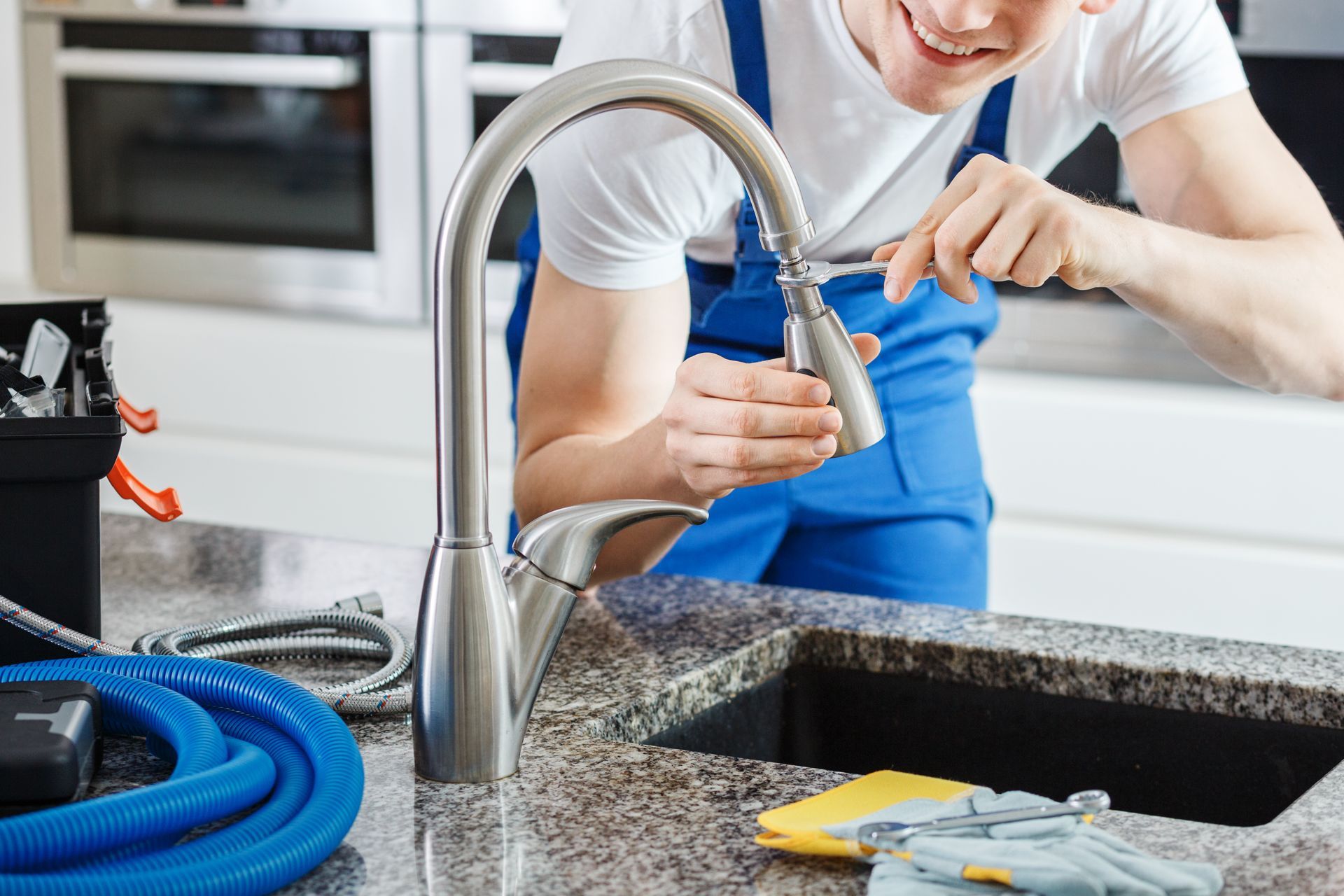 Plumber in blue overalls repairing a kitchen faucet with a screwdriver, smiling.