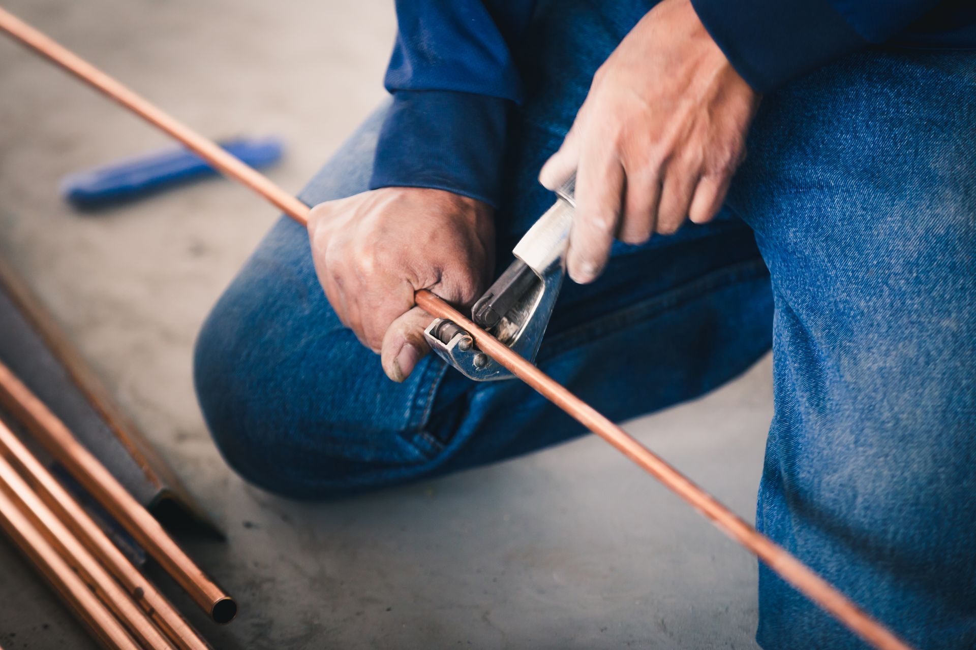 Person kneeling, cutting copper pipe with a pipe cutter. Blue jeans and shirt.