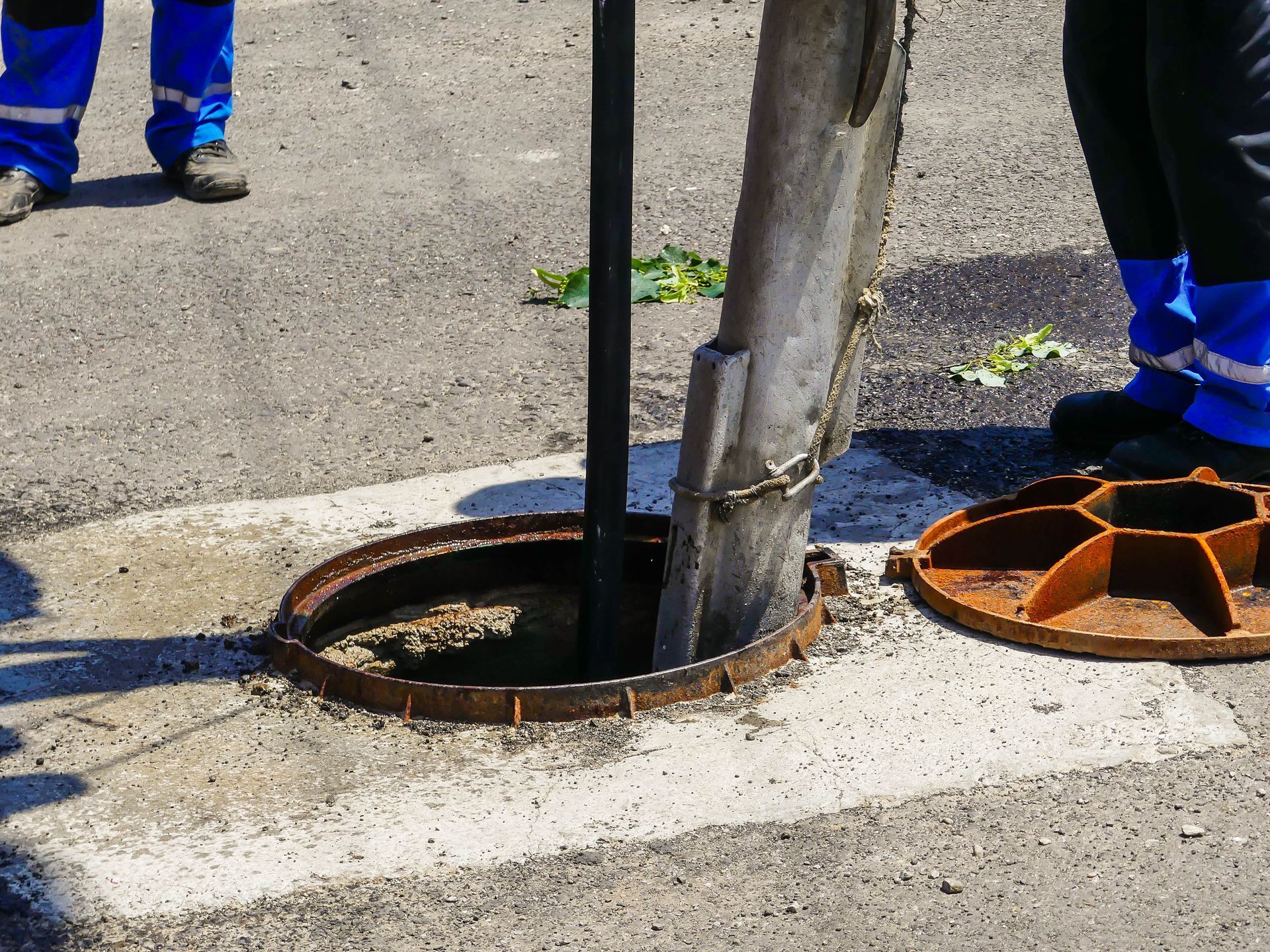 Workers inspecting a sewer with the manhole cover open on asphalt.