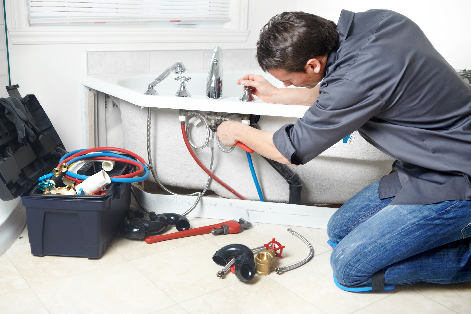 Plumber kneeling, working on pipes under a bathtub. Toolbox and tools are nearby.